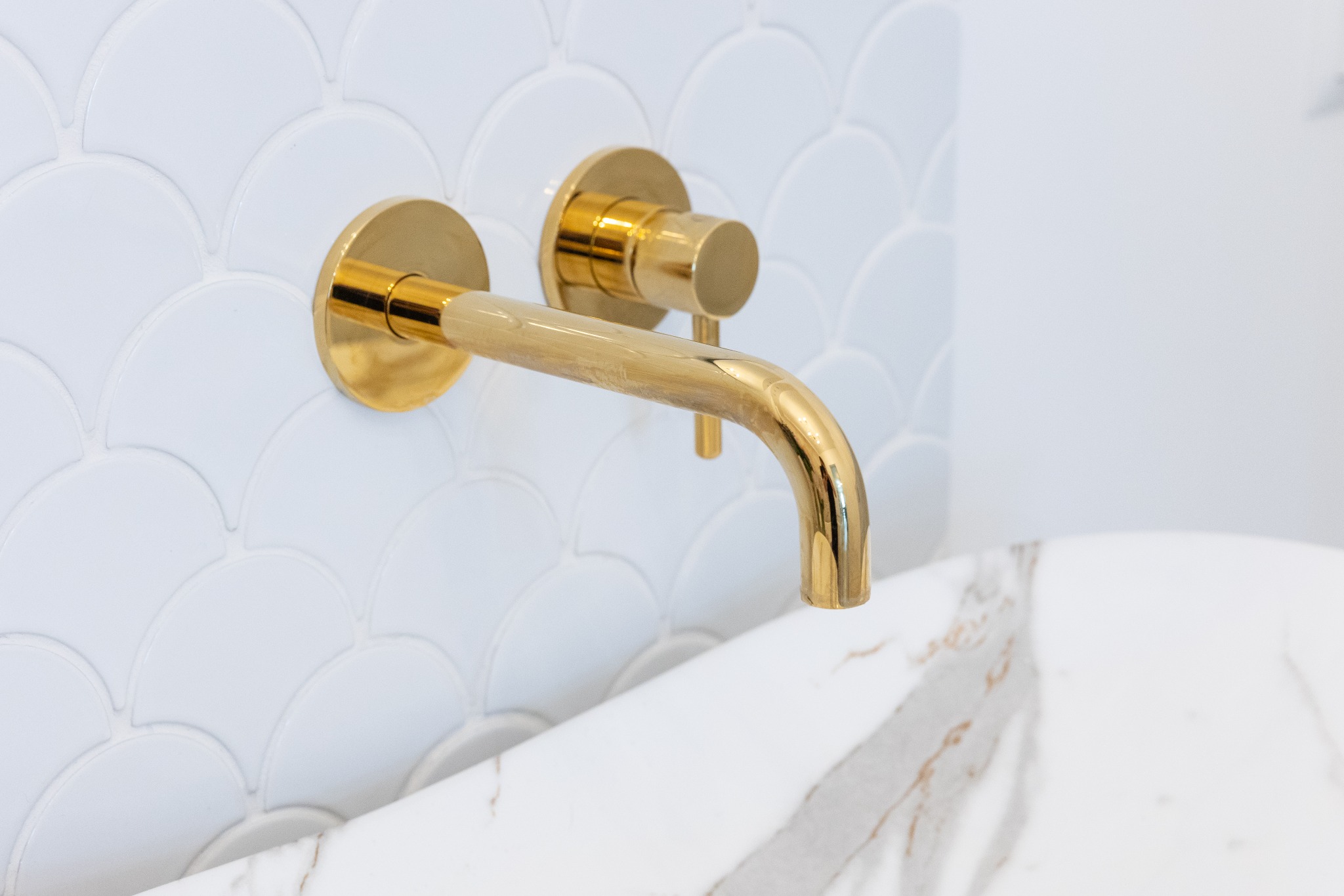 Bateria umywalkowa w kolorze złotym, zamontowana na ścianie z białych płytek w kształcie rybiej łuski, nad umywalką z białego marmuru.