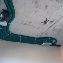 Zielone karbowane rury wentylacyjne przymocowane do surowego betonowego sufitu za pomocą metalowych taśm, widoczny fragment instalacji elektrycznej.