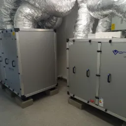 Dwa szare moduły centrali wentylacyjnej Vena Optima, połączone srebrnymi, elastycznymi przewodami wentylacyjnymi, umieszczone na betonowych podstawach w pomieszczeniu technicznym.