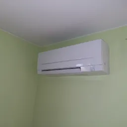 Biały klimatyzator Mitsubishi Electric zamontowany na jasnozielonej ścianie w rogu pokoju, z ozdobnym metalowym wieszakiem widocznym w prawym górnym rogu.