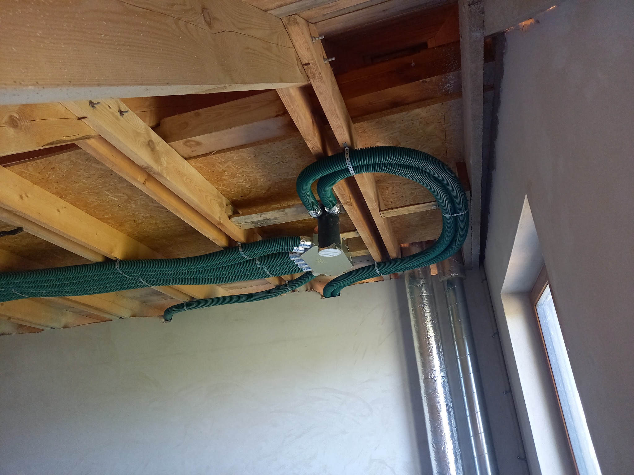 Zielone, karbowane rury wentylacyjne przymocowane do drewnianego stropu, widoczne srebrne kanały wentylacyjne w tle, surowy stan wykończenia ścian.