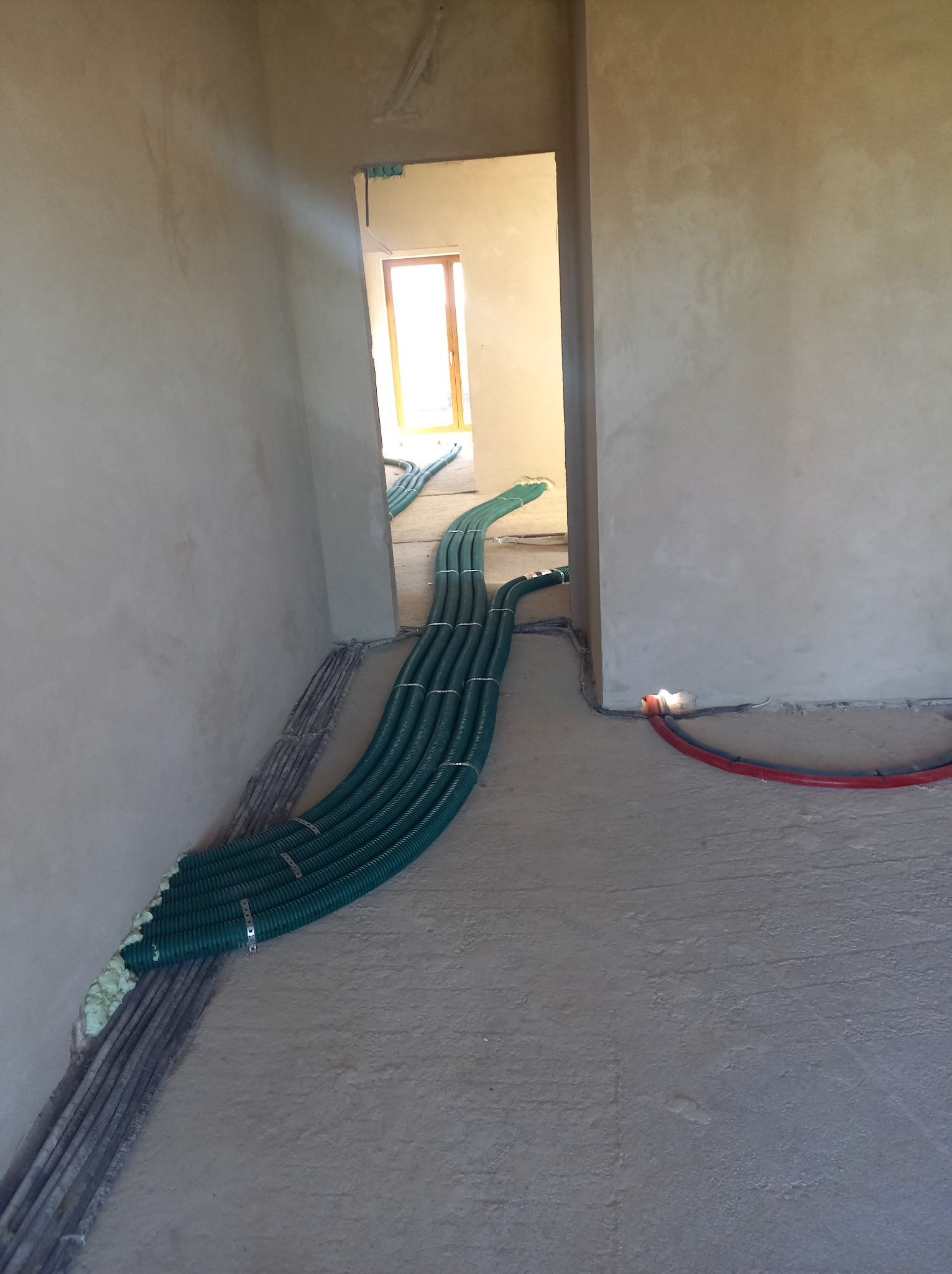 Zielone rury wentylacyjne ułożone na podłodze w surowym stanie, prowadzące przez próg drzwiowy, widoczne przygotowania do instalacji systemu wentylacji.