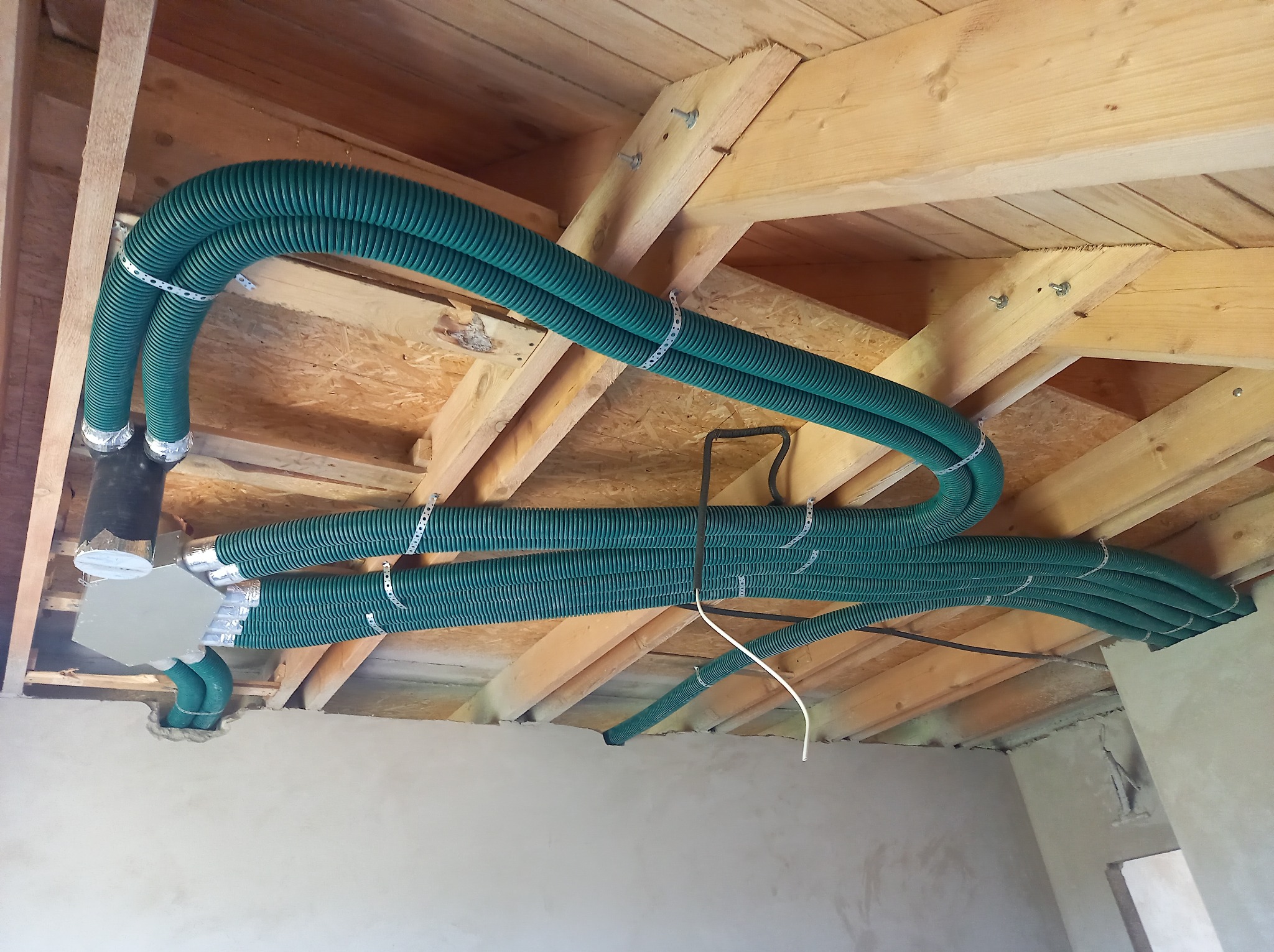 Zielone, karbowane rury wentylacyjne zamontowane pod drewnianym stropem, zabezpieczone metalowymi obejmami, widoczne elementy instalacji oraz surowy stan ścian.