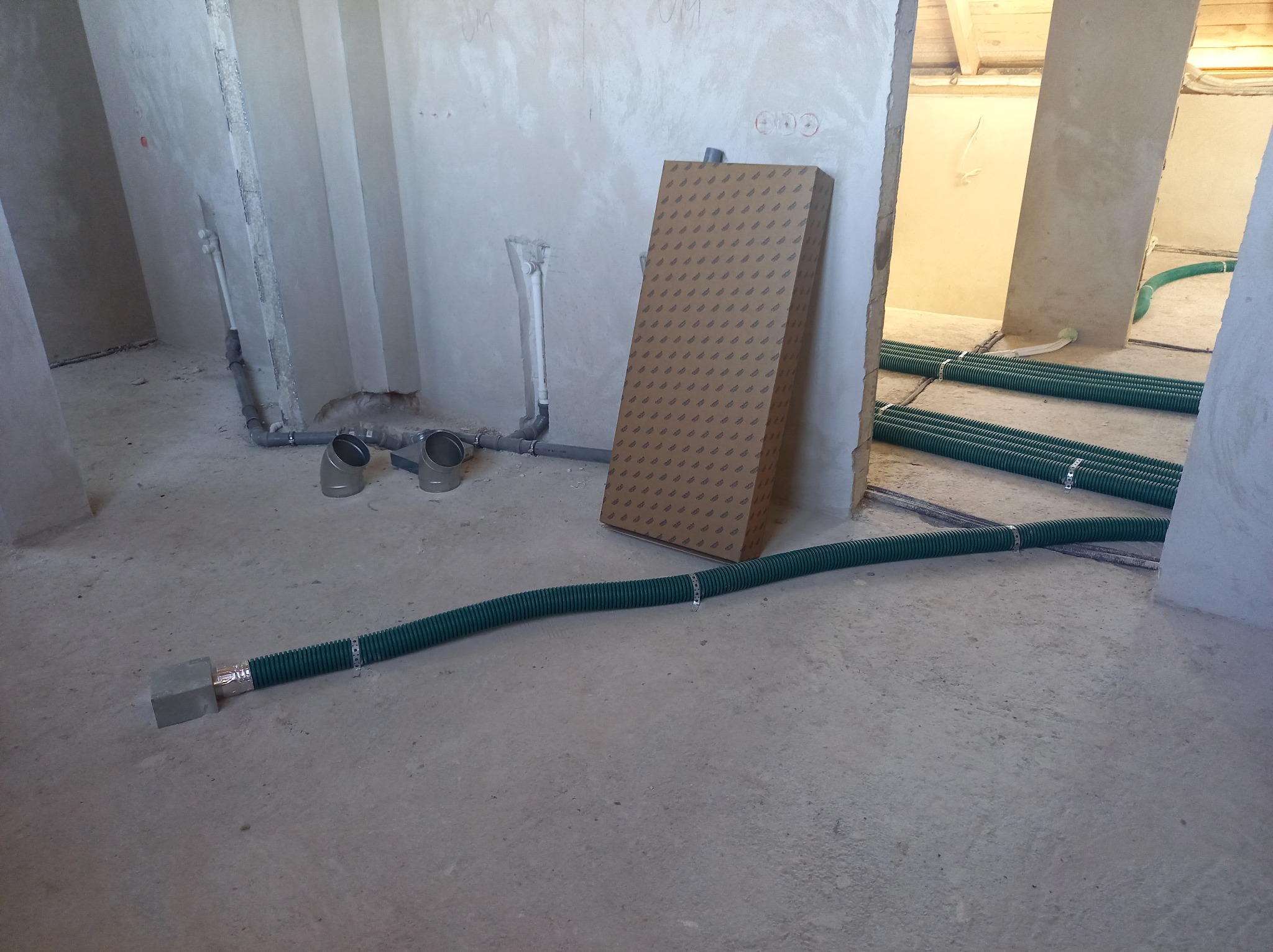 Wnętrze w stanie surowym z widocznymi rurami wentylacyjnymi w podłodze, szarymi rurami kanalizacyjnymi przy ścianie i kartonem zabezpieczającym.