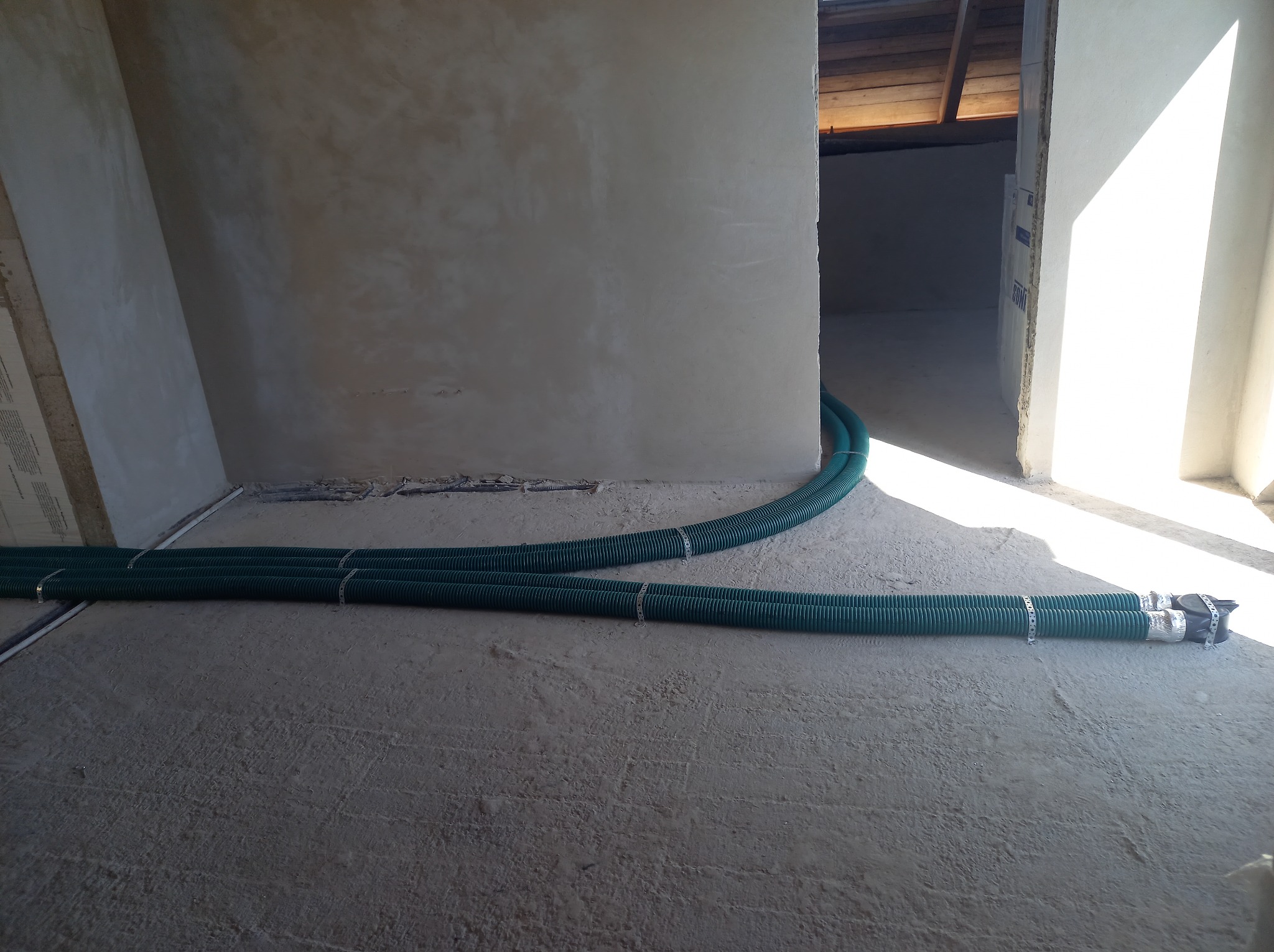 Trzy zielone, karbowane rury wentylacyjne z metalowymi opaskami leżące na surowym betonowym podłożu w trakcie instalacji w budynku w stanie deweloperskim, widoczne elementy konstrukcji dachu.