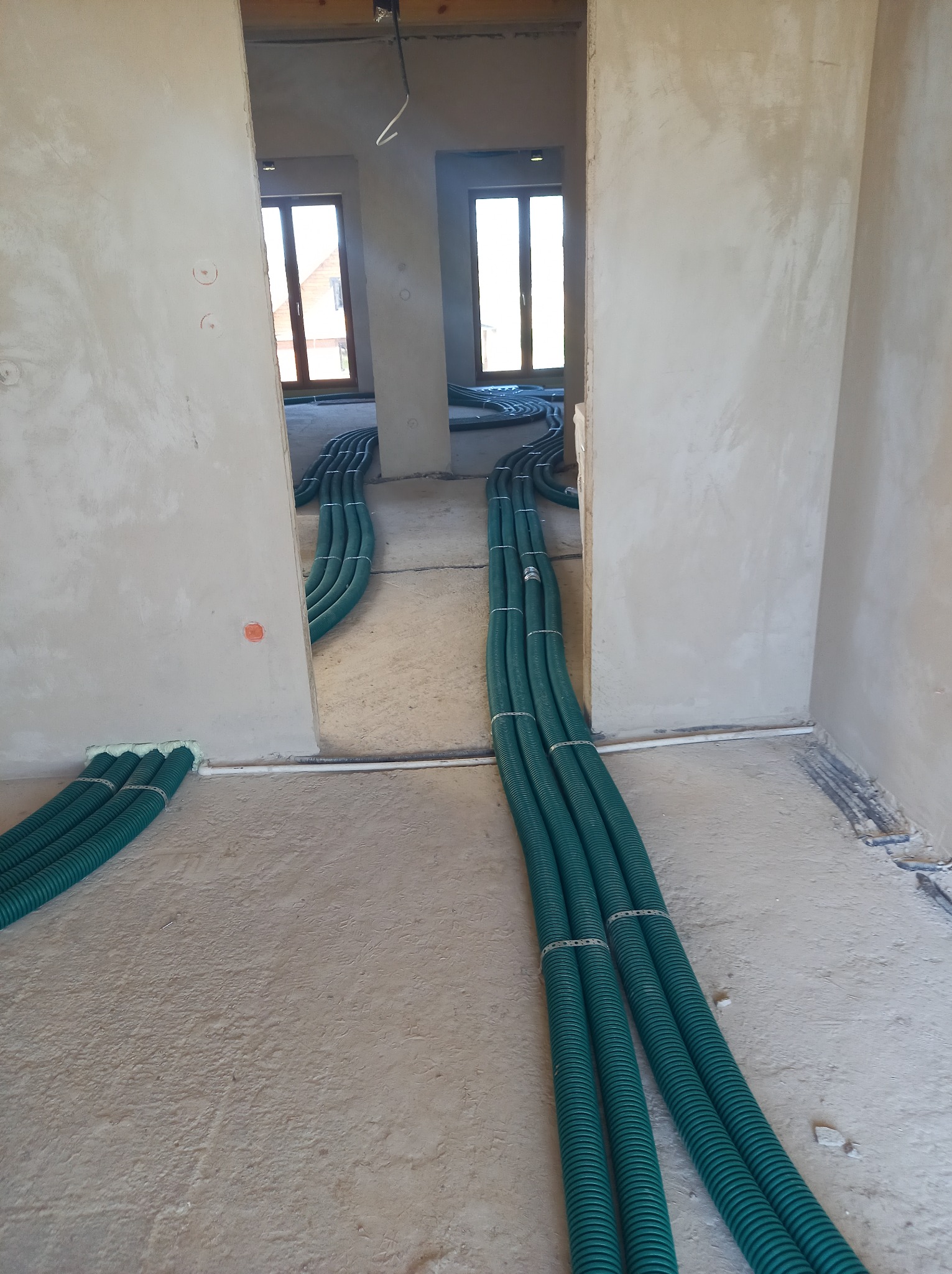 Wnętrze w stanie surowym z widocznymi, zielonymi rurami wentylacyjnymi ułożonymi wzdłuż podłogi i przechodzącymi przez otwory drzwiowe do kolejnych pomieszczeń.