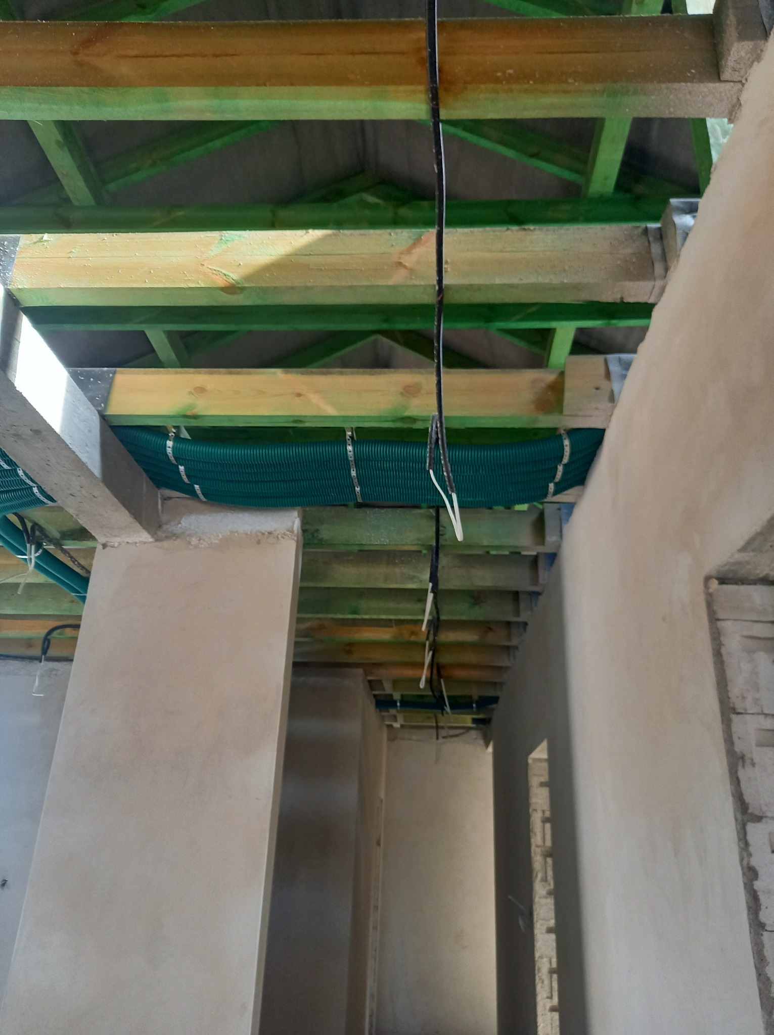 Wnętrze w trakcie budowy z widocznymi elementami instalacji wentylacyjnej w postaci zielonych rur karbowanych pod stropem drewnianym, przymocowanych metalowymi opaskami.