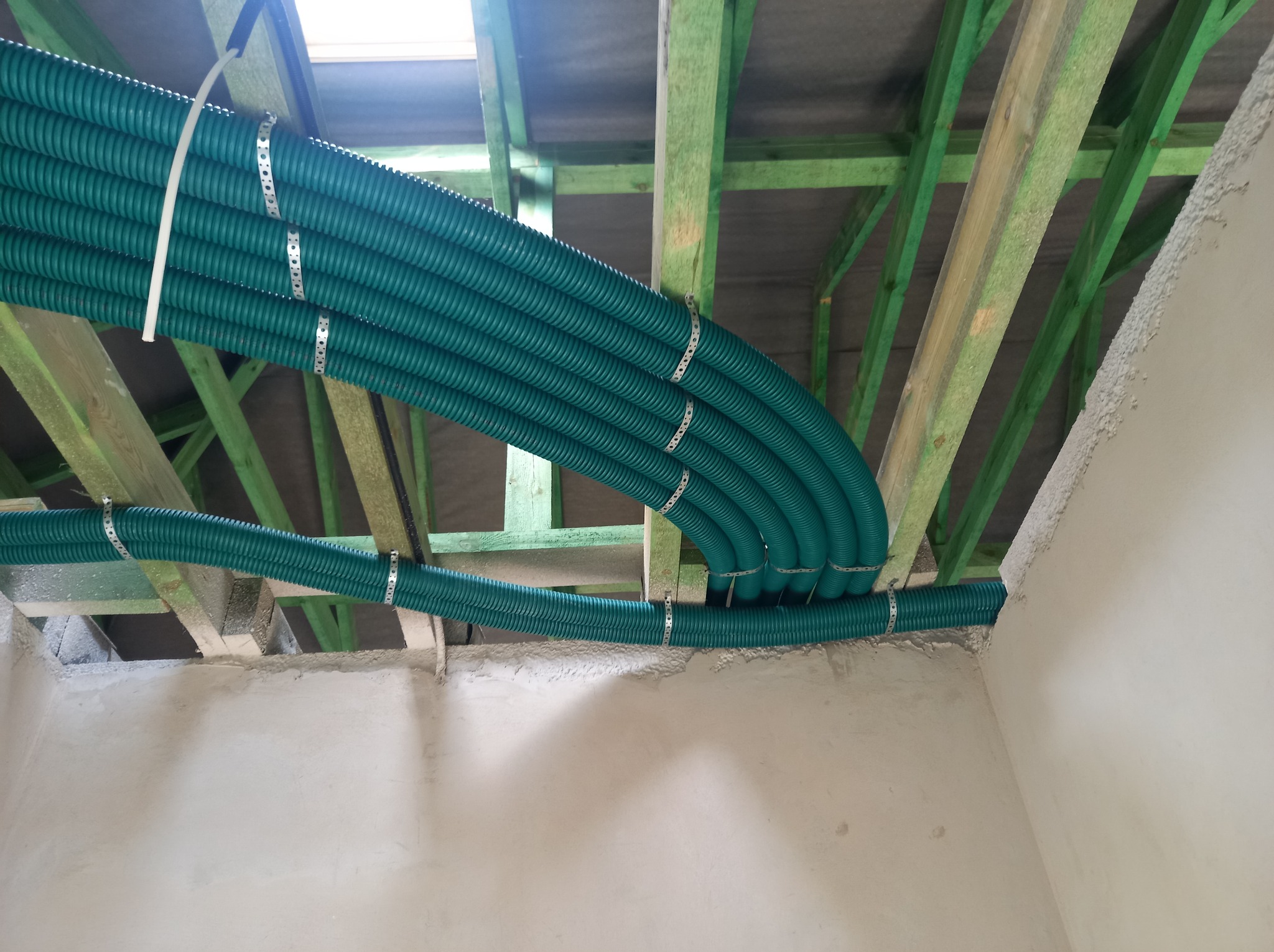 Szereg zielonych, karbowanych rur wentylacyjnych przymocowanych metalowymi obejmami do drewnianej konstrukcji stropu poddasza w surowym stanie.