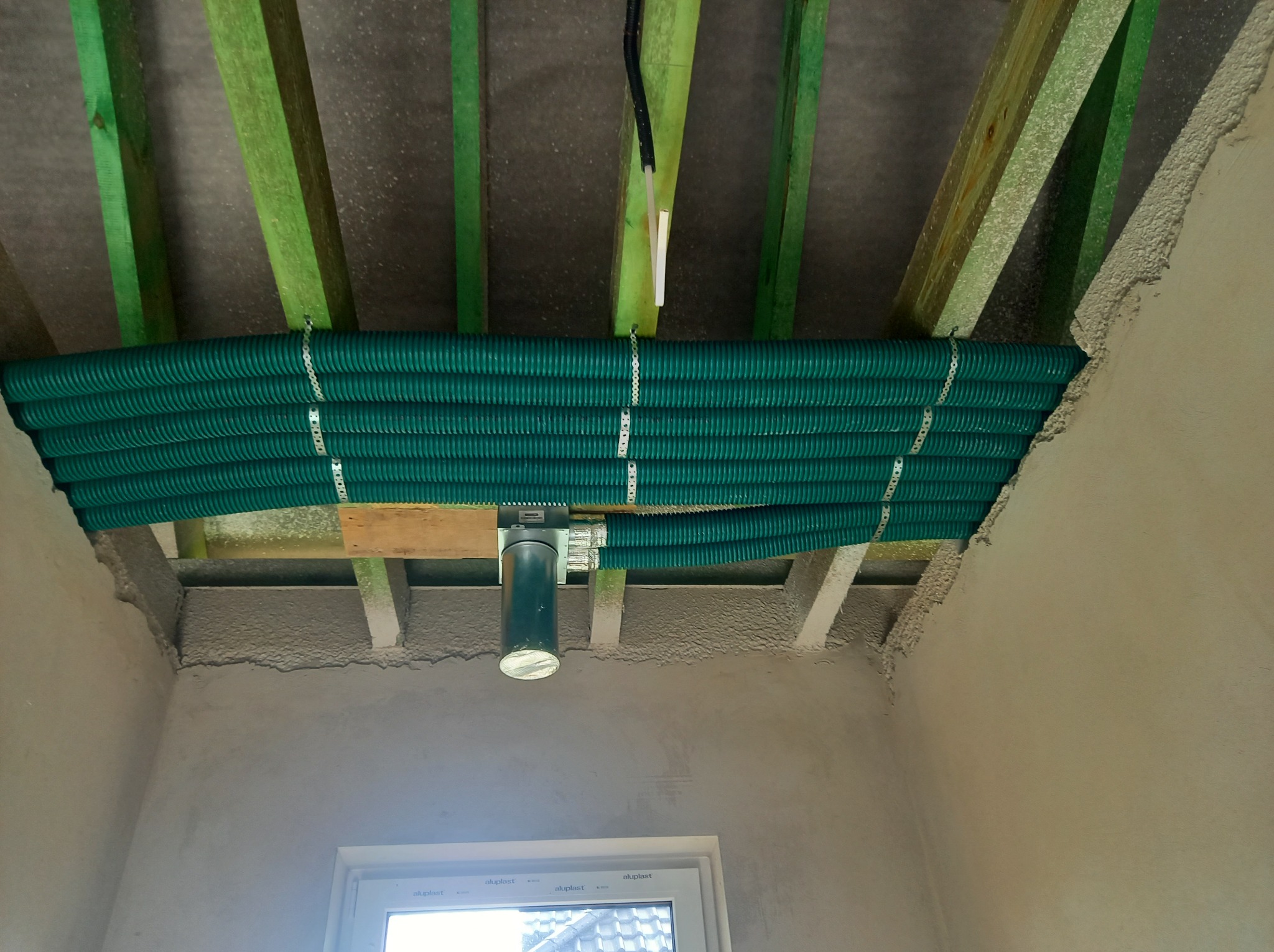 Szereg zielonych rur wentylacyjnych, przymocowanych metalowymi taśmami do drewnianej konstrukcji sufitu, z widocznym fragmentem metalowego kanału wentylacyjnego i surowymi ścianami w tle.