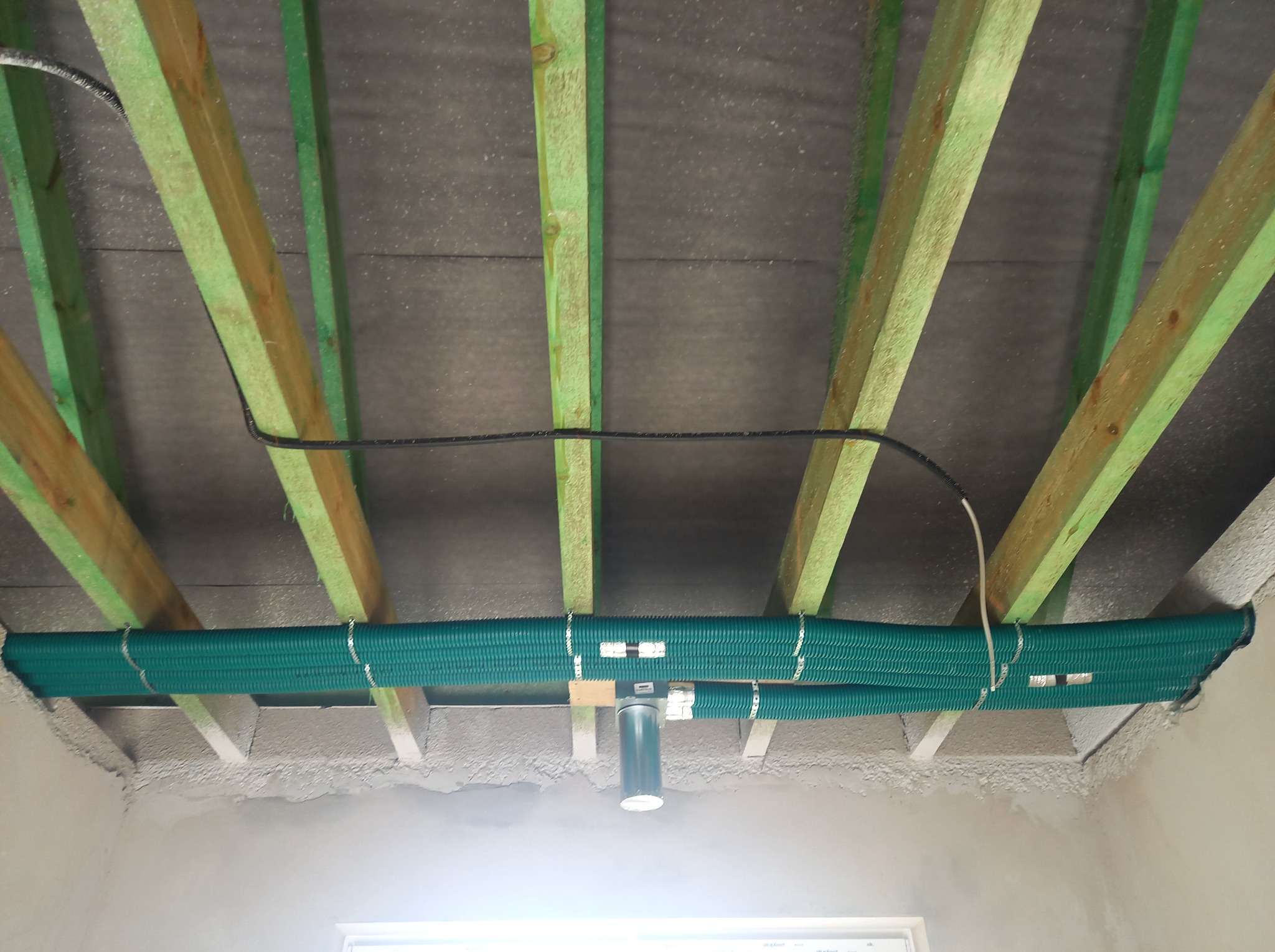Zielone rury wentylacyjne przymocowane do drewnianych belek stropowych z widoczną czarną instalacją elektryczną, w surowym stanie wykończenia wnętrza.