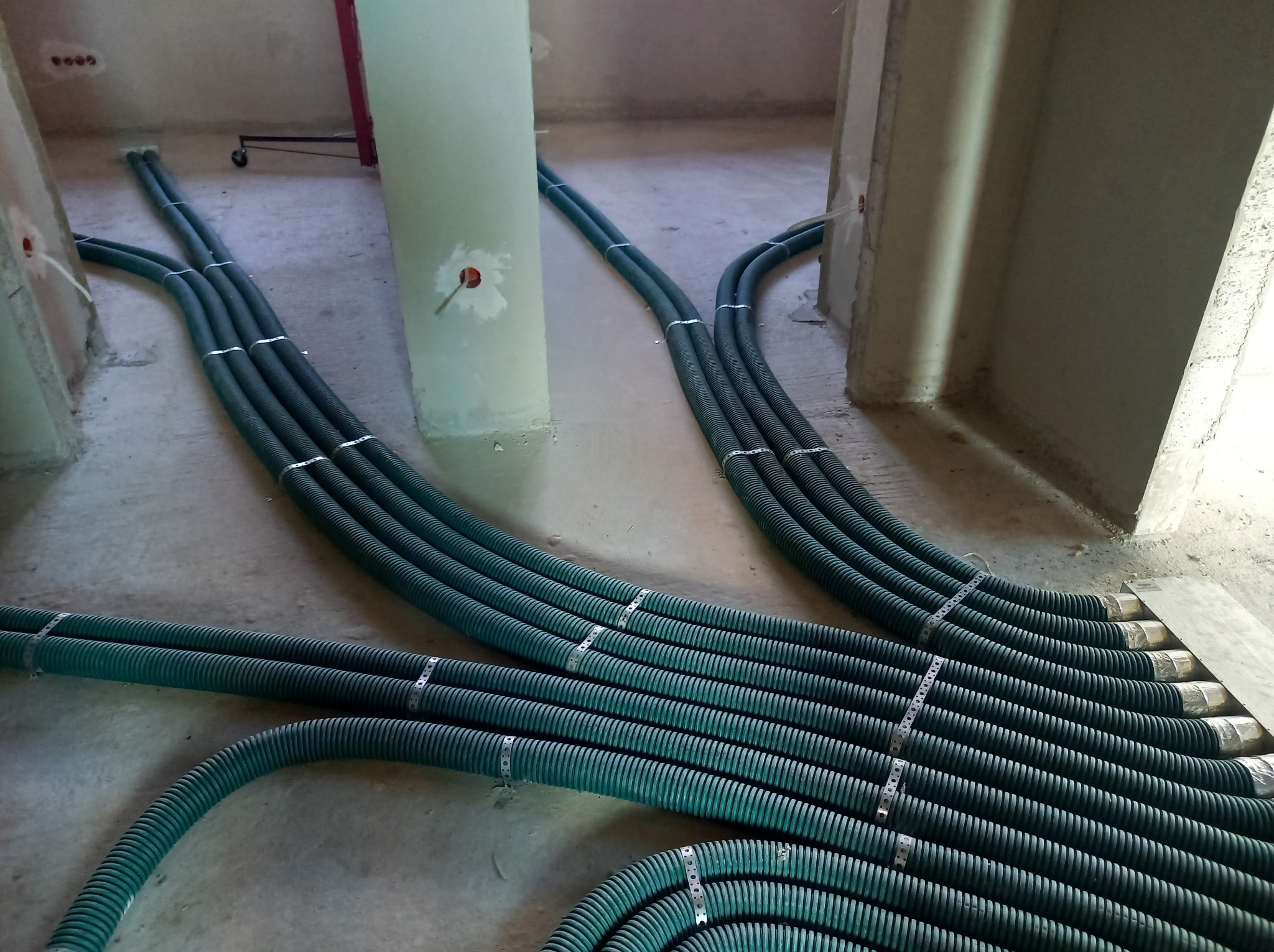 Zielone, karbowane rury wentylacyjne ułożone na surowej podłodze w trakcie montażu, zabezpieczone metalowymi opaskami, widoczne otwory w ścianach.