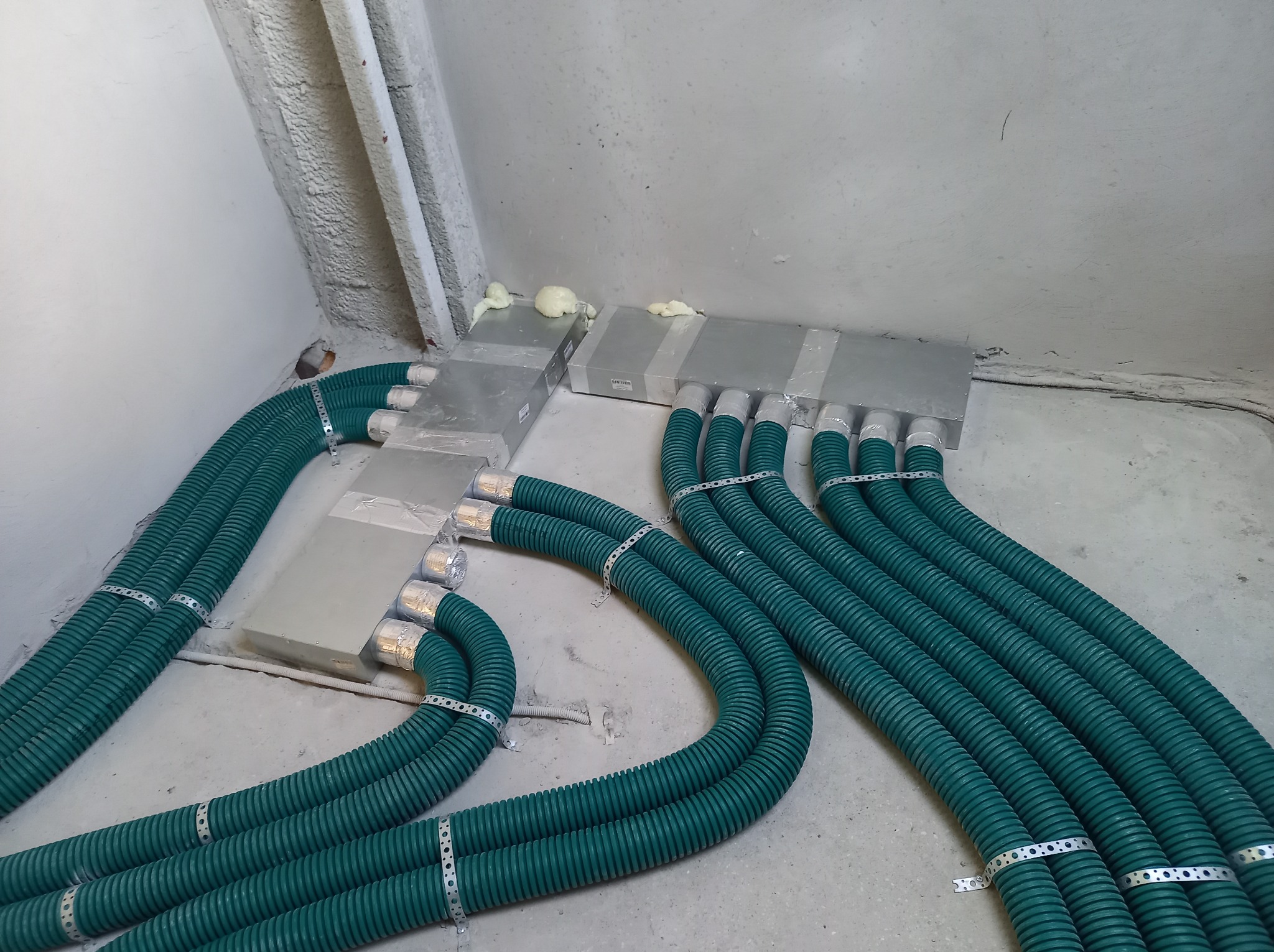 Szereg zielonych, karbowanych rur wentylacyjnych, połączonych srebrnymi skrzynkami i uszczelnionych pianką montażową, ułożonych na surowej posadzce w szarym pomieszczeniu.