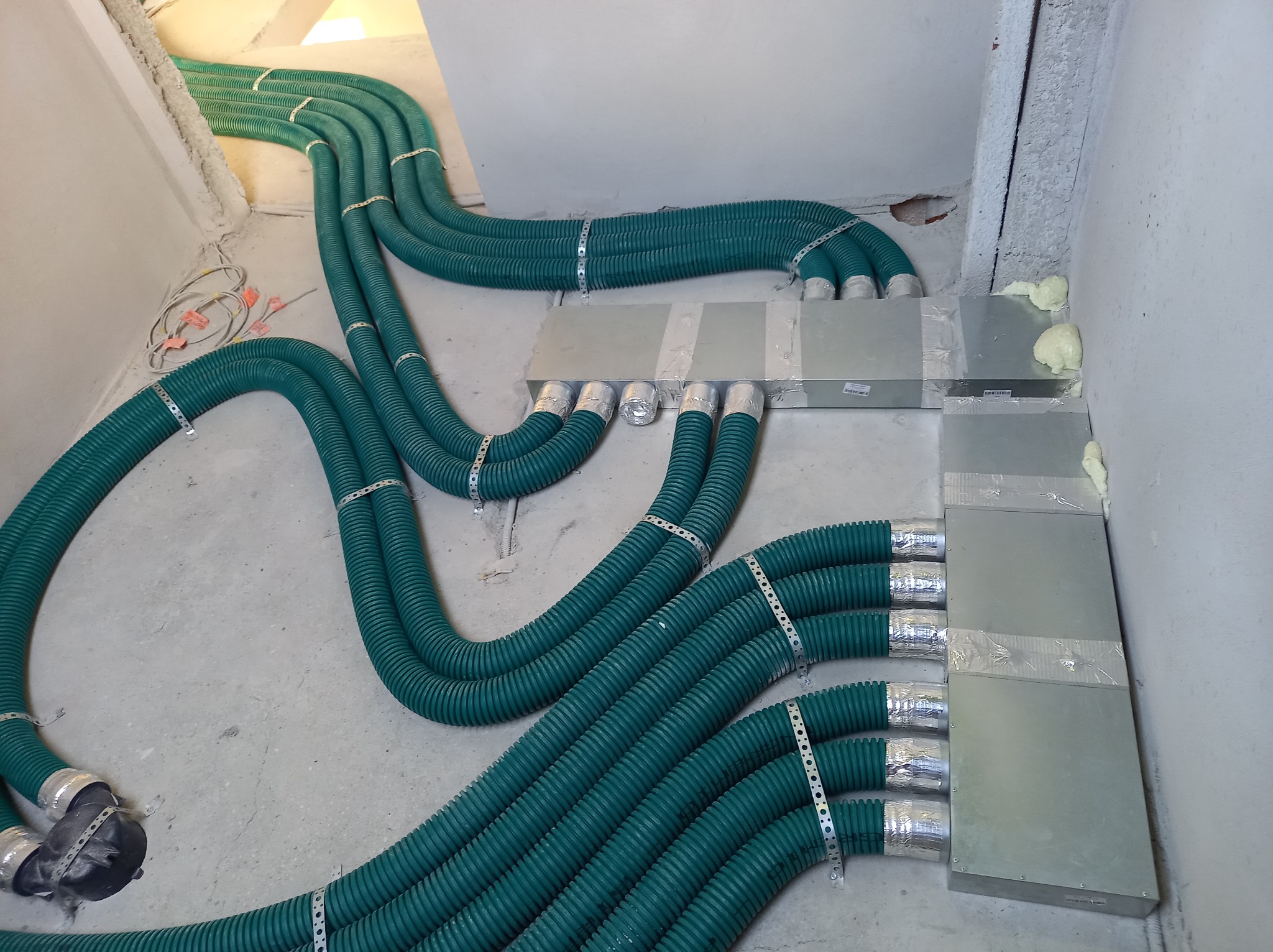 Zielone, karbowane przewody wentylacyjne przymocowane metalowymi opaskami, wpięte do metalowych skrzynek, uszczelnione pianką montażową przy ścianie.