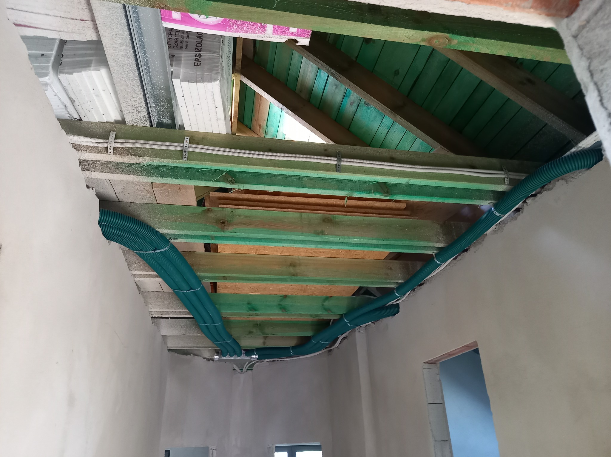 Zielone rury wentylacyjne prowadzone pod drewnianym stropem w surowym wnętrzu, widoczne materiały izolacyjne i częściowo otynkowane ściany.
