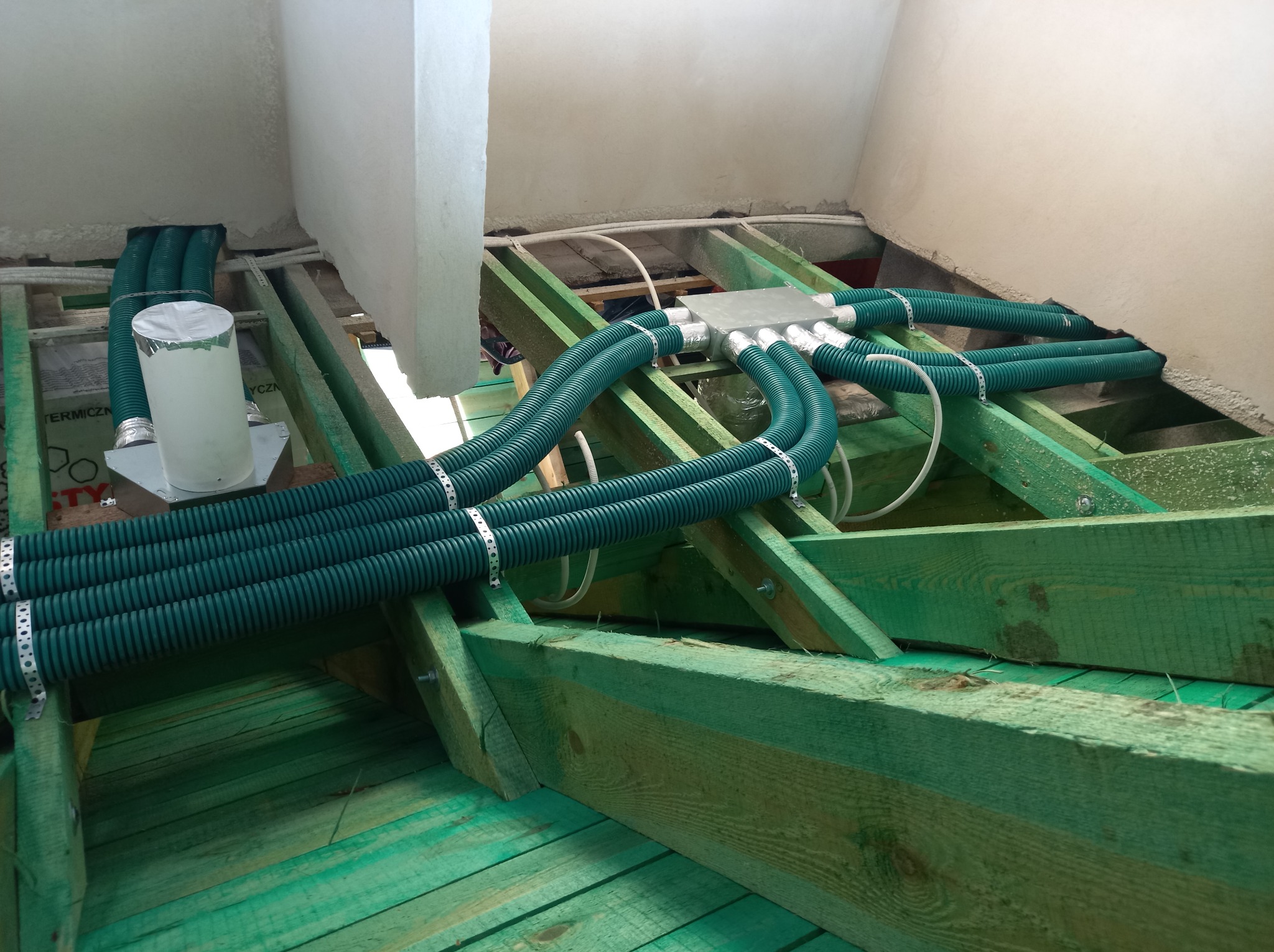 Zielone, karbowane rury wentylacyjne zamontowane na drewnianej konstrukcji stropu poddasza, połączone srebrną skrzynką rozdzielczą i przymocowane metalowymi opaskami.
