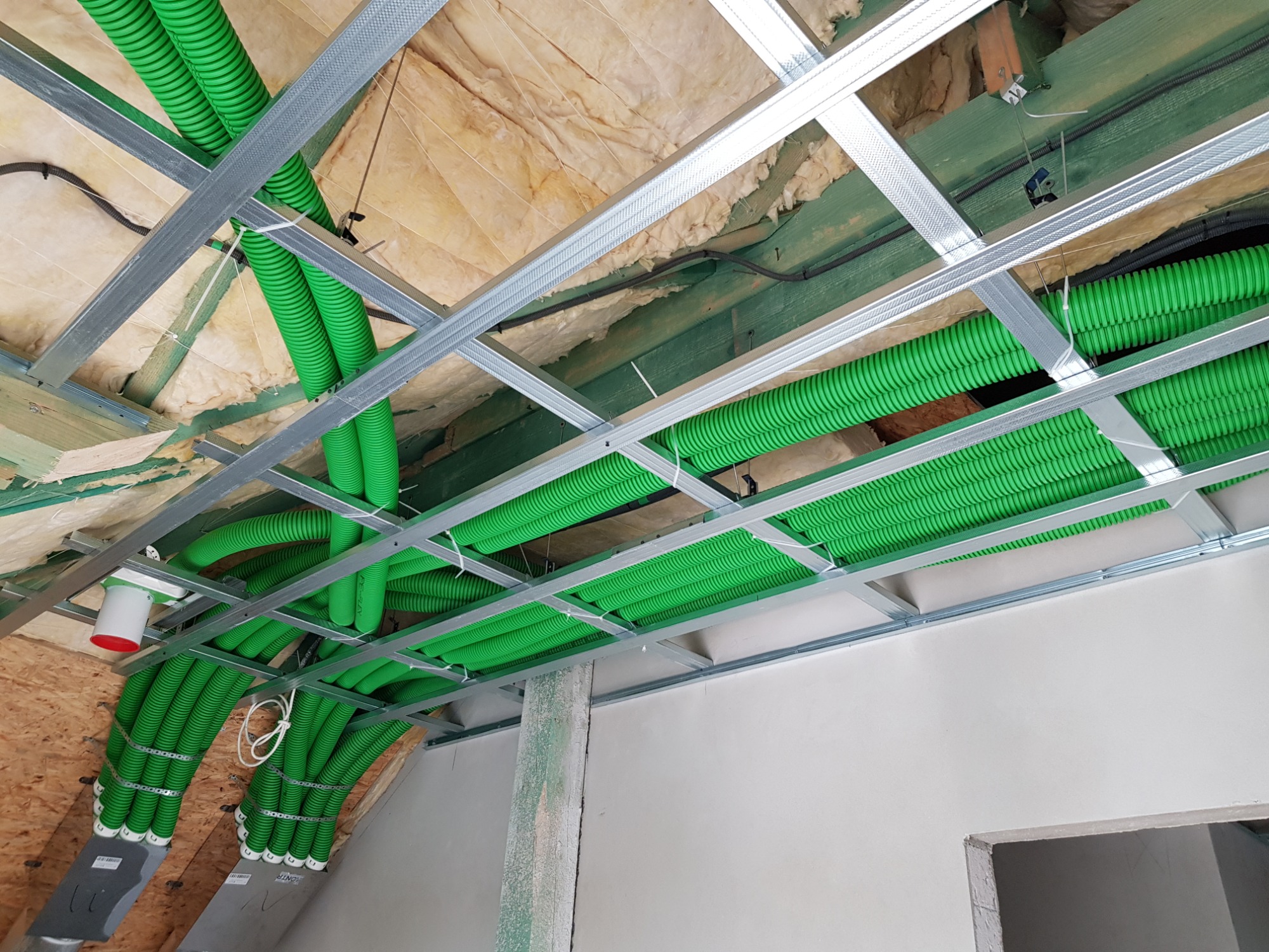 Zielone, karbowane rury wentylacyjne rozprowadzone pod sufitem podwieszanym z metalowej konstrukcji, widoczne ocieplenie z wełny mineralnej i elementy instalacji elektrycznej.