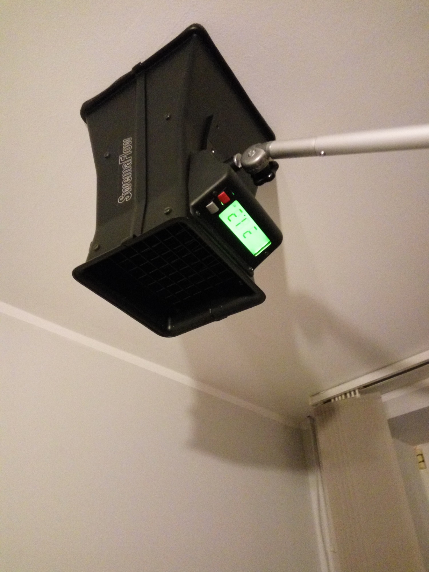 Czarny anemostat pomiarowy marki SwemaFlow zamocowany do sufitu na metalowym ramieniu, wyświetlający zielony odczyt 21.2.