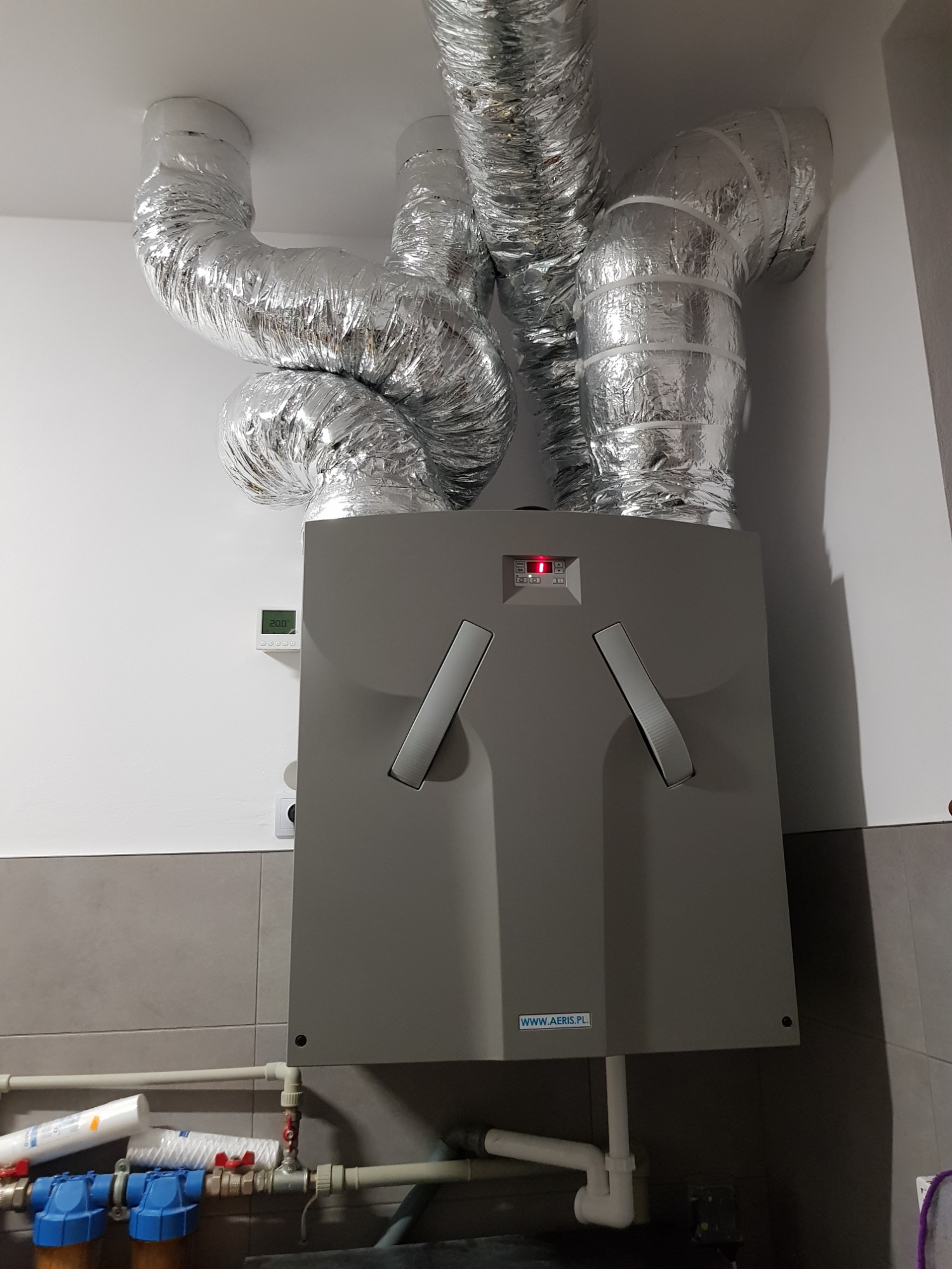 Szara centrala wentylacyjna z podłączonymi, elastycznymi przewodami aluminiowymi, zamontowana na ścianie z szarymi płytkami, widoczne filtry wody i rury.