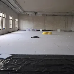 Przygotowanie podłoża pod wylewkę: rozłożone płyty izolacyjne na czarnej folii w surowym pomieszczeniu z dużymi oknami i widocznymi elementami instalacji pod sufitem.