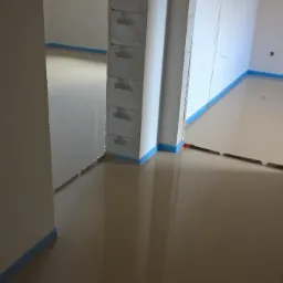 Świeżo wylana, gładka posadzka z widocznym niebieskim zabezpieczeniem krawędzi ścian. Widoczne ślady po łatach na łączeniu segmentów posadzki.