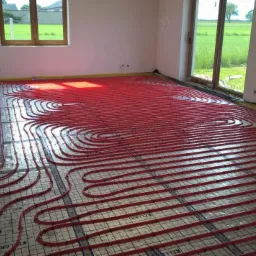 Czerwone rurki ogrzewania podłogowego ułożone na siatce w jasnym pomieszczeniu z widokiem na zielone pole przez okno.