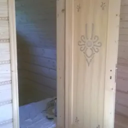 Drewniane drzwi wewnętrzne z dekoracyjnym, ażurowym wzorem kwiatowym, otwarte, widoczne drewniane wykończenie wnętrza w stylu góralskim.