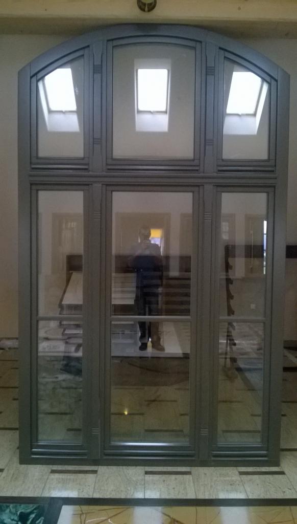 Szare, łukowe okno z trzema górnymi oknami dachowymi i trzema dolnymi szybami, odbicie osoby w środkowej szybie.