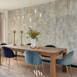 Jadalnia z drewnianym stołem, welurowymi krzesłami w odcieniach niebieskiego i złotymi nogami, złote świeczniki i nowoczesna lampa na tle ściany z teksturalną tapetą.