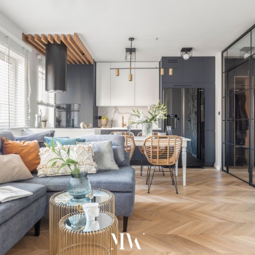 Jasne, nowoczesne wnętrze mieszkania z szarą sofą narożną, okrągłymi stolikami kawowymi ze złotymi nóżkami, parkietem w jodełkę, kuchnią w odcieniach szarości i bieli oraz jadalnią z białym stołem...