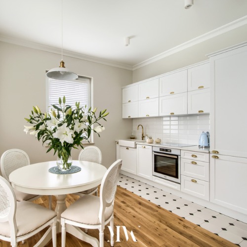 Jasna kuchnia z białymi meblami w stylu klasycznym, okrągły stół z bukietem białych lilii, drewniana podłoga przechodząca w czarno-białą mozaikę, złota armatura i błękitny czajnik.