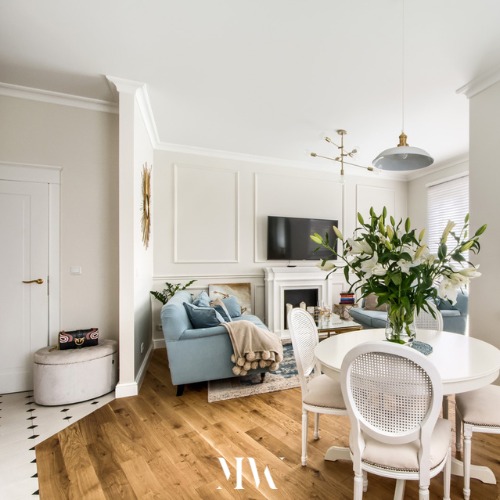 Jasne wnętrze salonu z jadalnią: błękitna sofa, biały stół z bukietem lilii, parkiet w jodełkę i dekoracyjne sztukaterie ścienne.