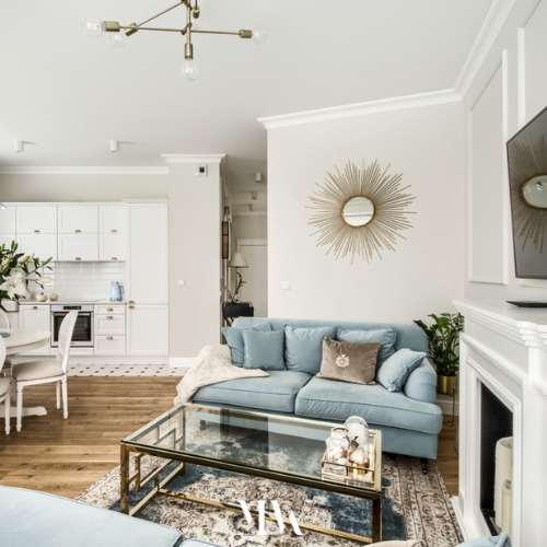 Jasne, przestronne wnętrze mieszkania z otwartą kuchnią, błękitną sofą, lustrem w kształcie słońca i szklanym stolikiem kawowym na wzorzystym dywanie.