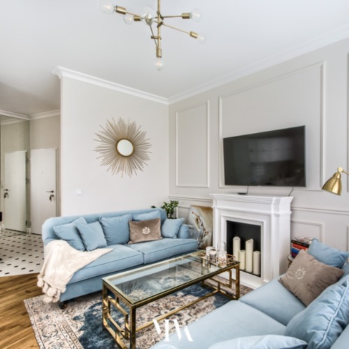 Jasne wnętrze salonu z błękitnymi sofami, szklanym stolikiem kawowym ze złotą ramą, kominkiem z białym obramowaniem i dekoracyjnym lustrem w kształcie słońca.