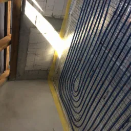 Niedokończona instalacja ogrzewania podłogowego z rurami grzewczymi ułożonymi na siatce, widoczne fragmenty surowego betonu i drewnianej konstrukcji dachu, oświetlone mocnym światłem słonecznym.