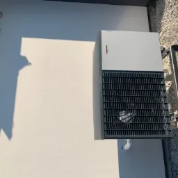 Szara jednostka zewnętrzna z czarną kratką wentylacyjną, zamontowana na ścianie budynku, widoczny fragment instalacji i podłoże z kamyczków.