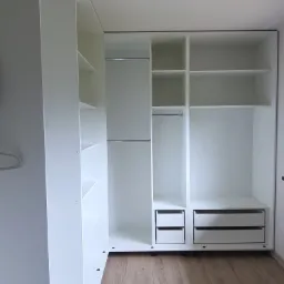 Wnętrze nowej, białej garderoby z półkami, drążkami i szufladami, widok z dołu na drewnianą podłogę, surowa żarówka zwisa z sufitu.