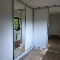 Wnętrze pokoju z zamontowaną szafą przesuwną z białymi frontami i lustrami, widoczne odbicie okna i grzejnika.