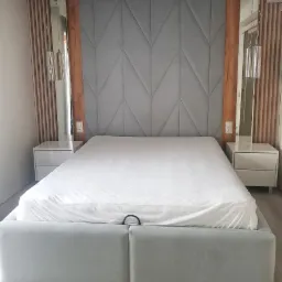 Szare, tapicerowane łóżko z geometrycznym wezgłowiem, biała pościel, szafki nocne z lampkami wiszącymi, drewniane panele dekoracyjne na ścianie.