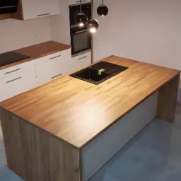 Drewniany blat wyspy kuchennej z czarnym zlewem, widok z góry, w tle białe szafki i wiszące lampy kuliste.