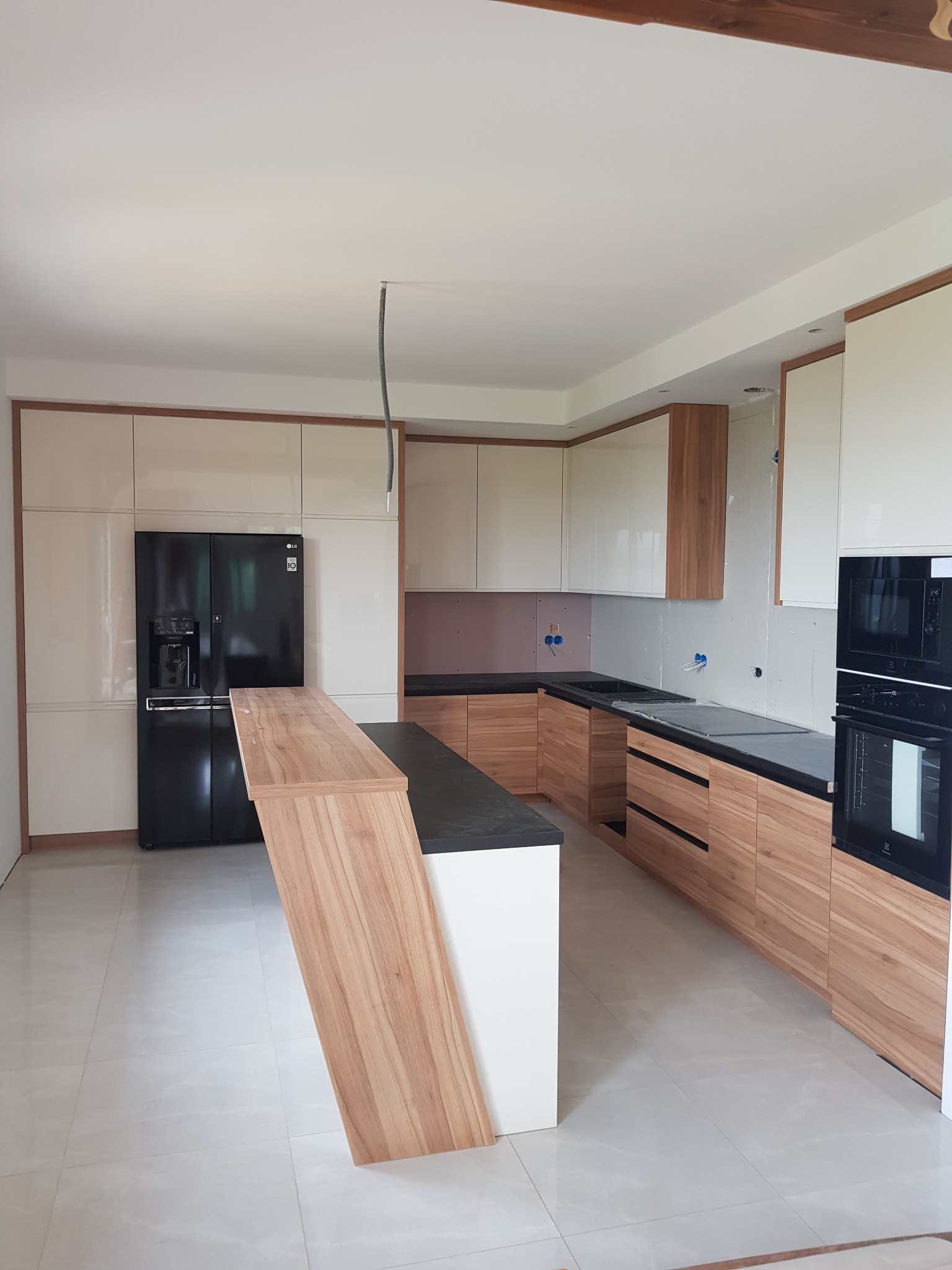 Nowoczesna kuchnia w trakcie montażu: jasne szafki górne z drewnianymi akcentami, drewniane szafki dolne z czarnym blatem, czarna lodówka side-by-side, wyspa z drewnianym blatem, widoczne...