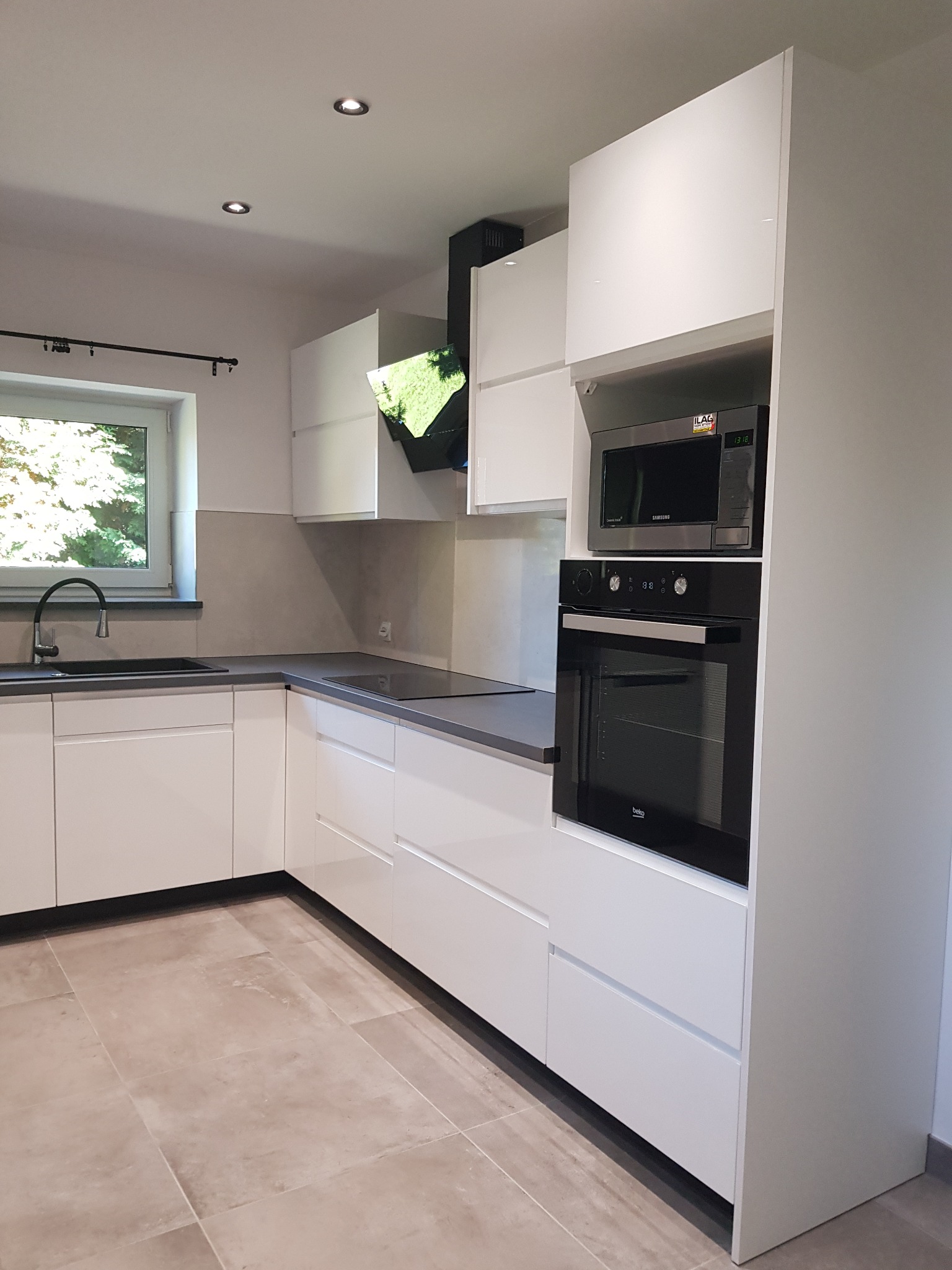 Nowoczesna, biała kuchnia z szarym blatem, czarną armaturą i sprzętem AGD w zabudowie, widok na okno z zielenią.