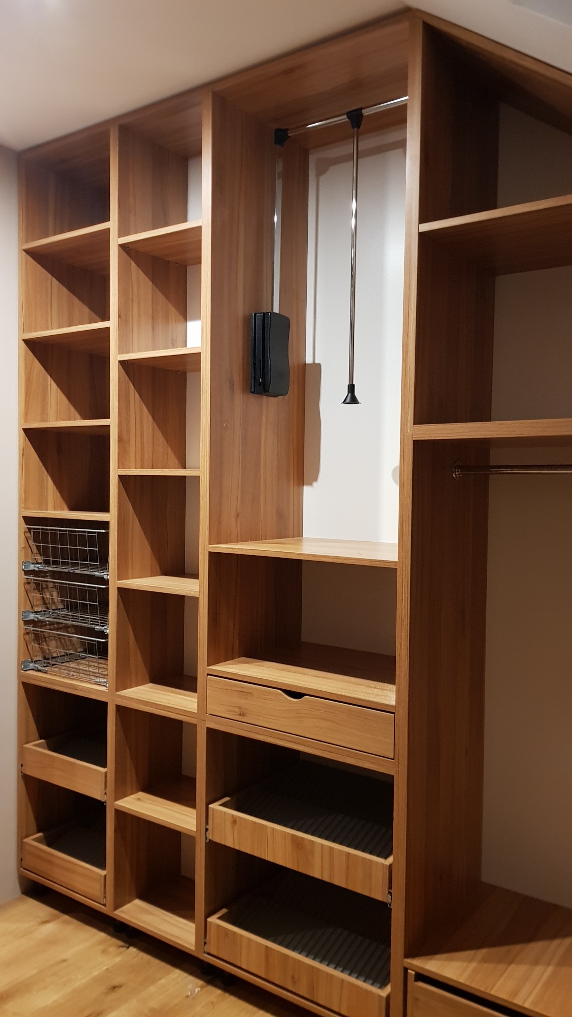 Drewniana garderoba z półkami, szufladami, koszami z siatki i drążkiem na ubrania, oświetlenie wewnątrz szafy, widoczny fragment drewnianej podłogi.