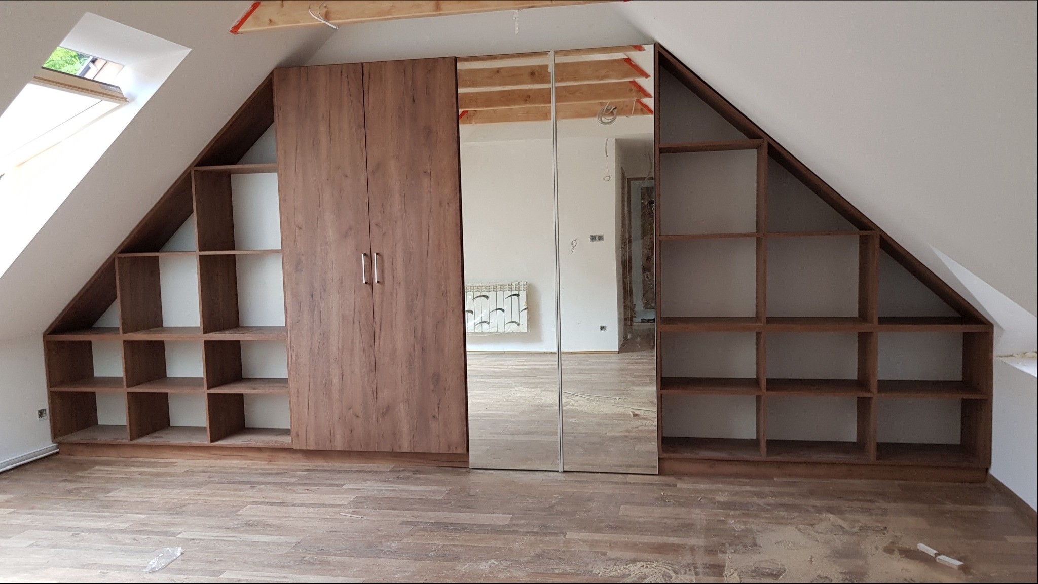 Zabudowa pod skosem na poddaszu z szafką z dwojgiem drzwi i lustrami, półki otwarte po bokach, drewnopodobny dekor, widoczne elementy instalacji na suficie.