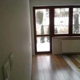 Układanie paneli podłogowych w jasnym wnętrzu z oknem na zimowy ogród, widoczne białe włączniki na ścianie.