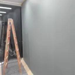 Świeżo pomalowana szara ściana z drewnianą drabiną w pomieszczeniu. Widoczne zabezpieczenia taśmą malarską przy podłodze i suficie.