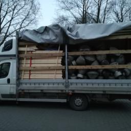 Carolus Magnus Sp. z o.o - Ciężarówka z otwartą skrzynią ładunkową, zabezpieczona plandeką, przewożąca drewniane belki i metalowe elementy w kształcie stożków, zaparkowana na ulicy.