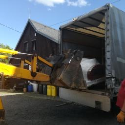 Carolus Magnus Sp. z o.o - Żółta ładowarka teleskopowa z łyżką umieszcza zapakowany w folię element w otwartej naczepie ciężarówki; w tle budynek z ciemnego drewna i pojemniki na śmieci.