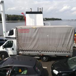 Carolus Magnus Sp. z o.o - Biała ciężarówka z szarą plandeką z napisem 'TRANSPORT PRZEPROWADZKI tel. 798-250-570' na promie, w tle woda i drzewa.