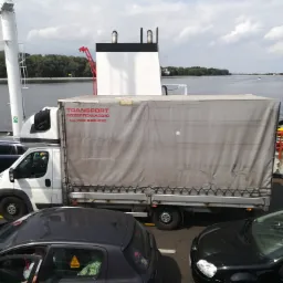 Biała ciężarówka z szarą plandeką z napisem 'TRANSPORT PRZEPROWADZKI tel. 798-250-570' na promie, w tle woda i drzewa.