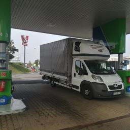 Carolus Magnus Sp. z o.o - Biały samochód dostawczy z plandeką stoi na stacji benzynowej OMV, obok dystrybutora z niebieską konewką i czerwoną gaśnicą, w tle widoczny szyld KFC.